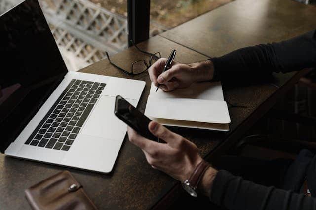 Person writing while using laptop and phone